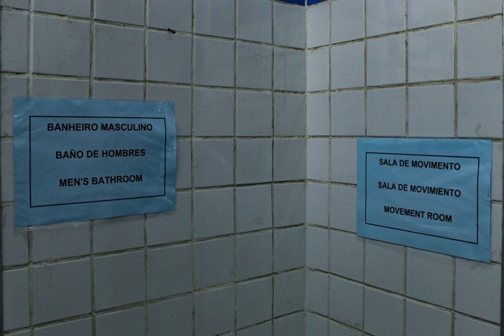 No banheiro masculino, a placa informa o local com os idiomas espanhol e inglês para os alunos imigrantes